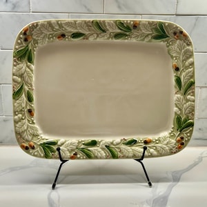 May include: Rectangular ceramic serving platter with a cream-colored center and a decorative border of green leaves, white foliage, and small orange accents. The platter is displayed on a black metal stand.