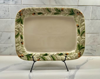 May include: Rectangular ceramic serving platter with a cream-colored center and a decorative border of green leaves, white foliage, and small orange accents. The platter is displayed on a black metal stand.