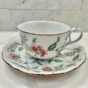May include: White teacup and saucer set with a floral design in pink, red, green, and blue. The cup and saucer have a gold trim. The teacup has a handle and sits on a matching saucer.