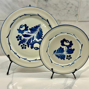 May include: Two white ceramic plates with blue floral designs and blue trim. The plates are displayed on black metal stands. The larger plate features a floral arrangement with leaves and berries. The smaller plate has a single flower and leaf design.