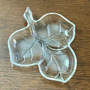 May include: A clear glass leaf-shaped serving dish with three sections. The dish has a stem and detailed vein patterns. The clear glass allows the contents to be visible. The dish is approximately 10 inches long.