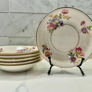 May include: A set of vintage cream-colored porcelain bowls and saucers. The bowls are stacked, and the saucers are decorated with colorful floral patterns and gold rims. One saucer is displayed on a black stand.