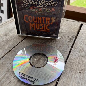 May include: A Great Ladies of Country Music CD in a clear plastic jewel case with a denim-textured cover featuring floral embroidery. The disc is silver with a holographic sheen, displaying the RCA Special Products logo and track information.