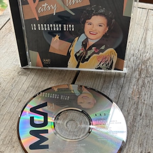 May include: A Patsy Cline 12 Greatest Hits compact disc in a clear plastic jewel case with a cover featuring a portrait of the artist, and a matching disc placed in front, displaying the MCA logo, track list, and Decca original catalogue text.