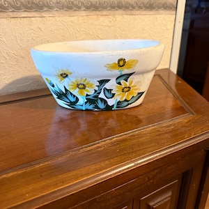 May include: White ceramic planter with a floral design. The planter has a rounded oval shape and features yellow flowers with green leaves painted on the side.