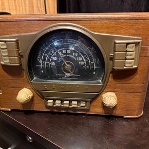 May include: A vintage Zenith tabletop radio with a rectangular wooden cabinet, a large circular glass dial featuring frequency markings, and a metallic faceplate. It includes multiple rectangular push buttons and two round knobs for tuning.