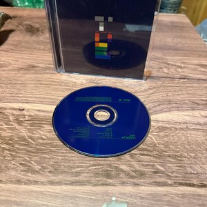 May include: A CD case and disc featuring a dark blue cover with a pixelated design in various colors. The CD is dark blue with text and a silver center. The case is open, revealing the disc inside.
