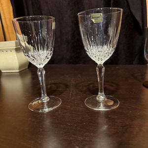 May include: Two clear crystal wine glasses with a decorative pattern on the bowl and stem. The glasses are on a dark brown surface. One glass has a small label attached. The glasses are empty.