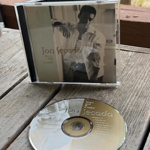 May include: A Jon Secada Heart, Soul & a Voice music CD in a clear plastic jewel case with a cover featuring a photograph of the artist. The disc itself is gold-toned with a tracklist and the artist's name printed in black text on the surface.
