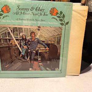 May include: A vintage vinyl record album by Sonny & Cher, titled "All I Ever Need Is You." The album cover is light green with orange floral accents and features a photo of the musical duo. The record is visible next to the album cover.
