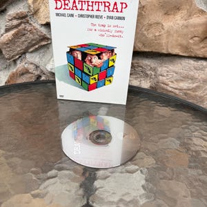 Peut inclure: Un boîtier DVD du film "Deathtrap" avec Michael Caine et Christopher Reeve. Le boîtier blanc affiche le titre en rouge. Un disque DVD est visible à côté du boîtier.