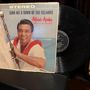 Peut inclure: Pochette d'album vinyle vintage intitulée "Sing Me a Song of the Islands" d'Alfred Apaka. La couverture présente un homme souriant portant un lei rouge, avec le logo Decca Records. Un disque vinyle noir est à côté de l'album.