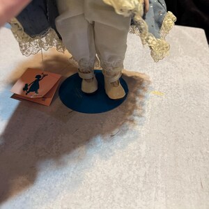May include: A porcelain doll with a blue dress and white pants. The doll has a tag with a silhouette of a girl holding flowers. The doll is standing on a blue base.