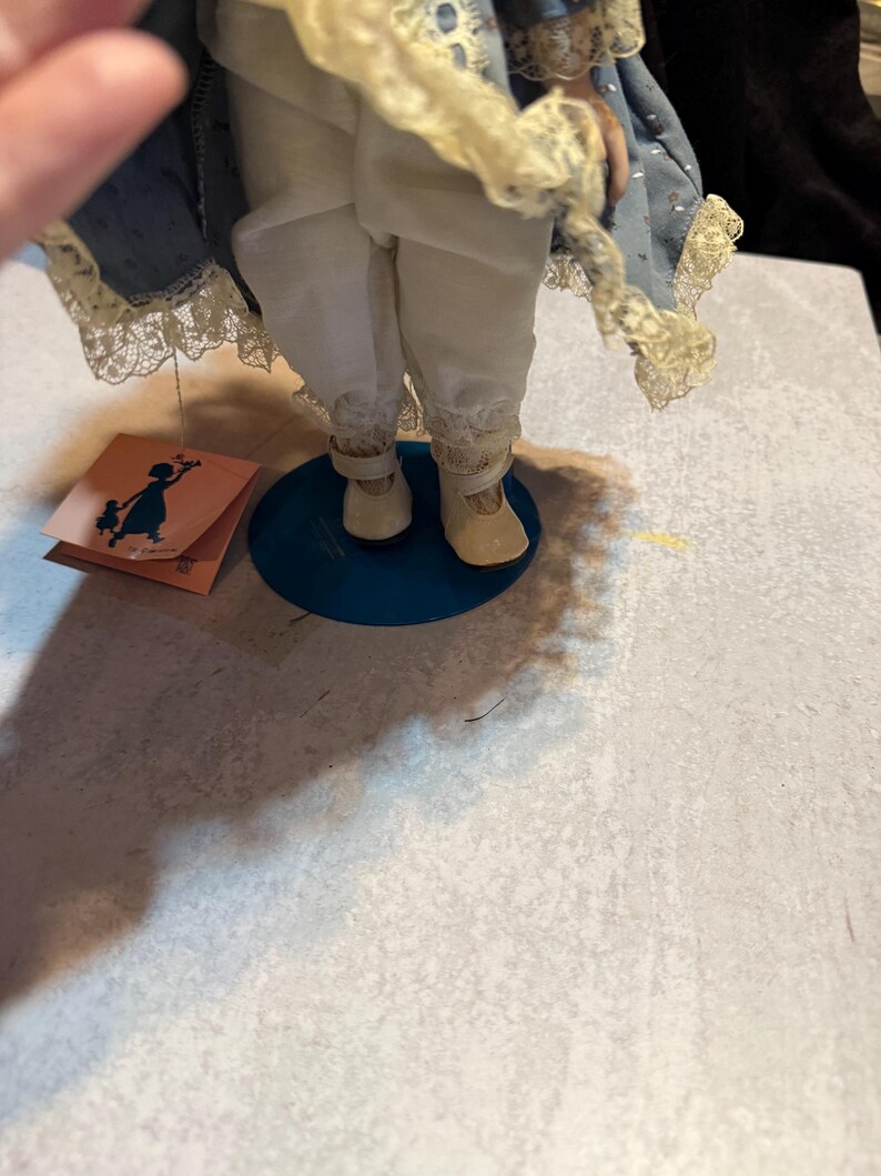 May include: A porcelain doll with a blue dress and white pants. The doll has a tag with a silhouette of a girl holding flowers. The doll is standing on a blue base.
