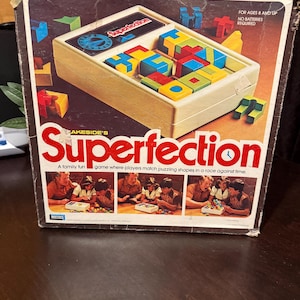 May include: Vintage Superfection game box. The box is brown with the game title in large red letters. The game features a beige plastic tray with colorful geometric shapes. Text on the box reads "A family fun game where players match puzzling shapes in a race against time."