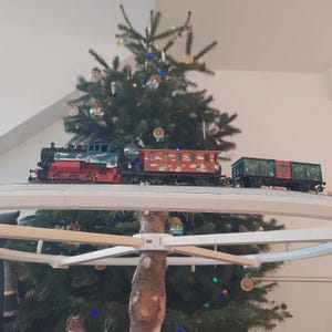 Könnte beinhalten: Eine Modelleisenbahn mit einer roten, schwarzen und blauen Lokomotive, die zwei Waggons zieht. Der Zug fährt auf einer Strecke vor einem geschmückten Weihnachtsbaum. Die Waggons sind rot und grün mit festlichen Designs.