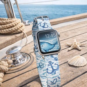 Könnte beinhalten: Eine silberne Smartwatch mit einem blauen Wal-Motiv auf dem Bildschirm und einem Armband im maritimen Stil mit Ankern und Wellen. Die Uhr zeigt "Tue 24 12:45". Die Uhr liegt auf einer Holzoberfläche mit nautischen Elementen.