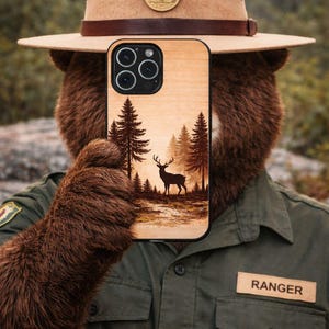 May include: A wooden phone case featuring a forest scene with a deer and pine trees. The case is held by a person wearing a ranger hat and uniform. The hat has a brown leather band and a gold emblem. The uniform has a "RANGER" patch.