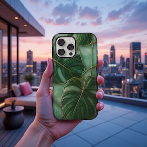Può includere: Una custodia per telefono verde e oro con un design a foglie di monstera. La custodia ha una finitura lucida e dettagli dorati. La custodia è tenuta davanti a uno skyline urbano al tramonto.