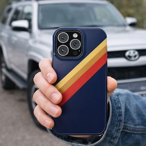Op de afbeelding: Een marineblauwe telefoonhoes met een diagonaal streepontwerp in geel, oranje en rood. De hoes heeft een vierkante cameramodule met meerdere lenzen. Op de achtergrond is een zilveren SUV te zien.