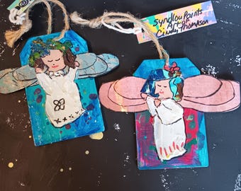 May include: Two handmade angel ornaments. Each angel has a white dress, blue hair, and pink wings. The ornaments are painted on wood with colorful backgrounds. One tag reads "@syndlouka" and the other reads "Syndlou Paints Art of Cindy Thompson."