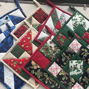 May include: A collection of handmade quilted potholders with various holiday-themed patterns. Designs include snowmen, poinsettias, holly, and plaid, in shades of blue, red, green, and white. Each potholder has a loop for hanging.