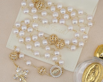 Puede incluir: Un rosario dorado con perlas blancas y detalles dorados ornamentados. El rosario presenta un colgante de cruz con piedras transparentes y un colgante circular con una figura religiosa. El rosario se exhibe sobre una bolsa blanca.