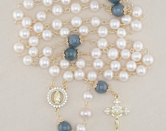 Puede incluir: Un rosario con perlas blancas, cuentas gris azuladas y detalles dorados. El collar incluye un colgante de crucifijo con cristales transparentes y un medallón con una figura religiosa.