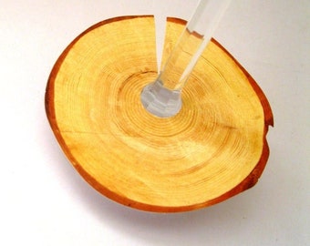 May include: A wooden coaster with a circular shape and a hole in the center. The coaster is made of light brown wood with darker brown rings. A clear glass wine glass is resting on the coaster.