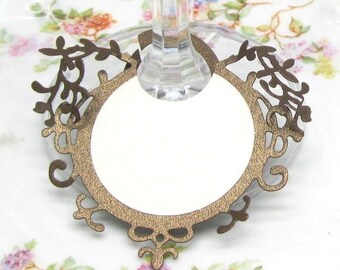 May include: A gold-colored metal frame with a circular cutout for a place card. The frame is designed with a delicate, intricate pattern of leaves and swirls. It is attached to a clear glass stem.