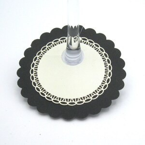 May include: A black and cream-colored drink coaster with a scalloped edge. The coaster has a cream-colored center with a decorative white lace-like border. A clear glass stem sits in the center of the coaster.