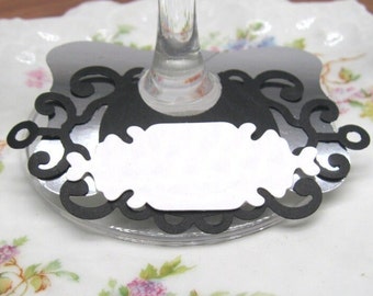 May include: Black and white paper wine glass charm with a decorative design and a blank space for writing a name.