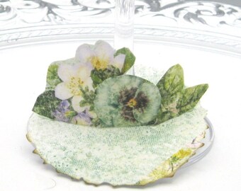 May include: A green and white paper doily with a floral design featuring pansies and other flowers. The doily is attached to a clear plastic base.