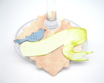 May include: A light pink and yellow paper wine glass charm with a blue bird and a blank banner. The charm is designed to hang from a wine glass.