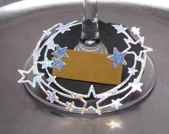May include: Silver glitter star-shaped wine glass charms with a blank card for a name. The charms are in the shape of a crescent moon.