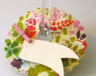 May include: A white card with a scalloped edge and a pink clothespin attached to the top. The card is attached to a glass stem with a floral patterned base.