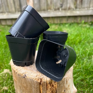 May include: Four black square plastic plant pots are stacked on a tree stump. The pots are empty and have drainage holes at the bottom. The background includes green grass and a wooden fence.