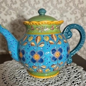 Könnte beinhalten: Eine blaue und grüne Teekanne aus Keramik mit floralem Muster. Die Teekanne hat einen gelben Rand und einen gewellten Deckel.