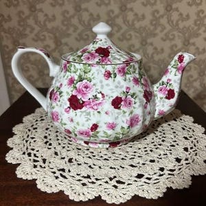Op de afbeelding: Een witte porseleinen theepot met een herhalend patroon van donkerrode en lichtroze rozen met groene bladeren. De theepot heeft een ronde vorm, een gebogen handvat, een bijpassend deksel met een puntige knop en een gedetailleerde tuit.