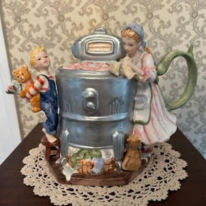 Puede incluir: Una tetera de cerámica con forma de lavadora vintage con una figura de mujer y un niño en la parte superior. La mujer sostiene una prenda de vestir y el niño sostiene un oso de peluche. La tetera está decorada con un acabado plateado y tiene un asa verde.
