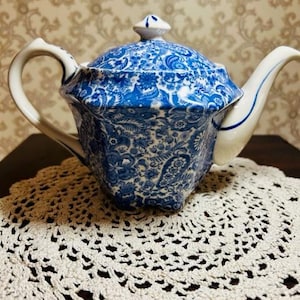 May include: A blue and white teapot with a floral pattern. The teapot is sitting on a white crocheted doily.