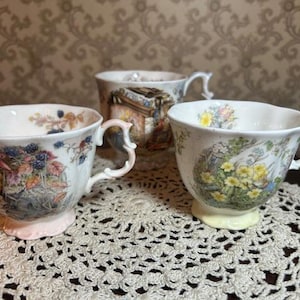 May include: Three white porcelain teacups with scalloped edges and ornate handles. Each cup features a distinct colorful botanical illustration, including blackberries, yellow flowers, and a woodland scene, all set on a white base with footed rims.