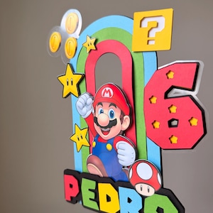 May include: A multi-layered cake topper featuring a cartoon character in a red hat and blue overalls, a large red number 6 with yellow stars, a yellow question mark block, gold coins, a red mushroom, and a rainbow arch with the name PEDRO.