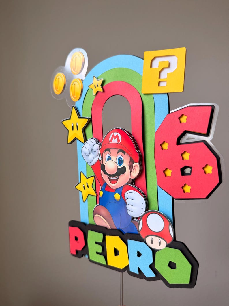 May include: A multi-layered cake topper featuring a cartoon character in a red hat and blue overalls, a large red number 6 with yellow stars, a yellow question mark block, gold coins, a red mushroom, and a rainbow arch with the name PEDRO.