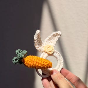 May include: A handmade crochet baby rattle in the shape of a bunny with a carrot. The bunny is white with yellow accents, and the carrot is orange with green leaves. The rattle is held in a hand.