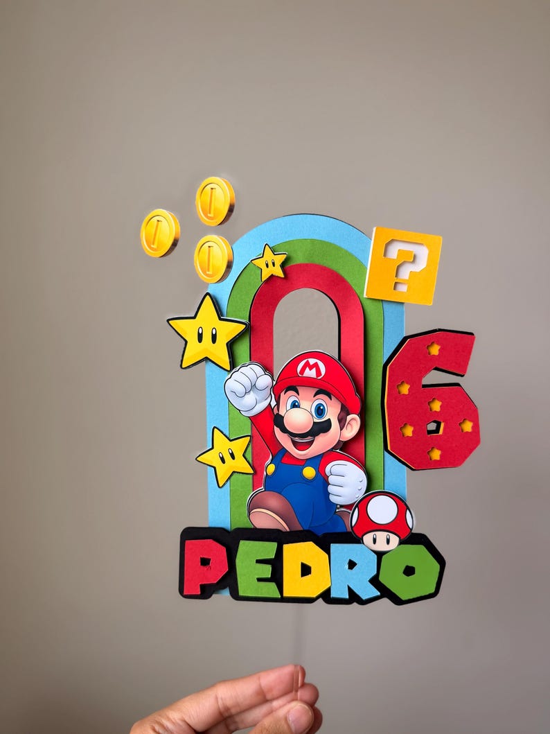 May include: A multi-layered cake topper featuring a cartoon character in a red hat and blue overalls, a rainbow arch, yellow stars, gold coins, a yellow question block, a red mushroom, and the number 6, with the name PEDRO in colorful letters.