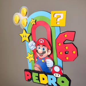 May include: A multi-layered cake topper featuring a colorful rainbow arch, a Mario character, a red number 6 with yellow stars, a yellow question mark block, gold coins, and a red mushroom. The name PEDRO is displayed in colorful block letters.