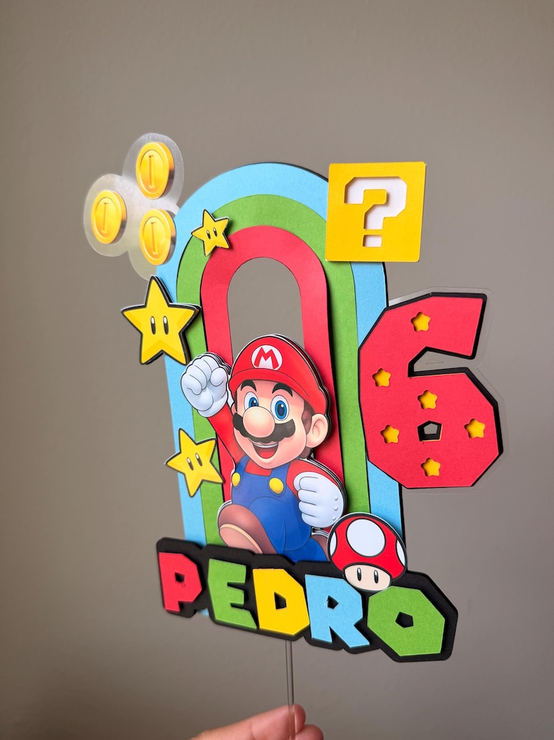 May include: A multi-layered cake topper featuring a colorful rainbow arch, a Mario character, a red number 6 with yellow stars, a yellow question mark block, gold coins, and a red mushroom. The name PEDRO is displayed in colorful block letters.