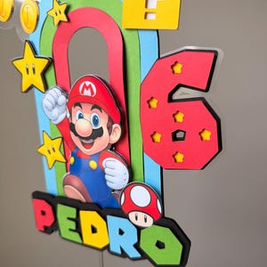 May include: A multi-layered cake topper featuring a colorful rainbow, a red number six with star cutouts, and a character wearing a red hat and blue overalls. The name PEDRO is displayed in block letters, accompanied by a mushroom and gold coins.