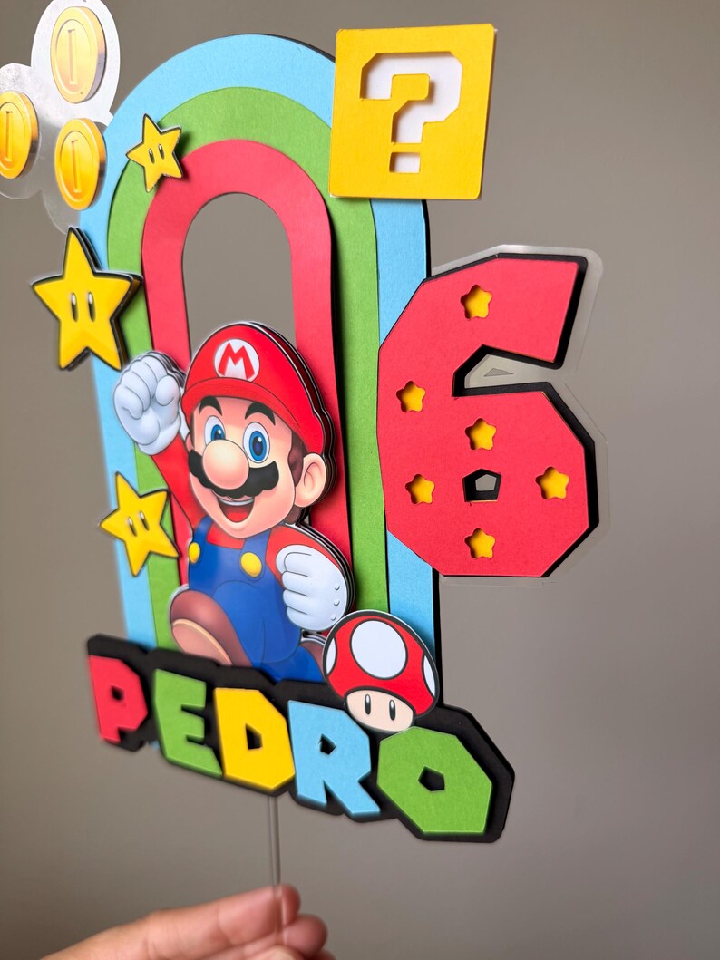 May include: A multi-layered cake topper featuring a colorful rainbow, a red number six with star cutouts, and a character wearing a red hat and blue overalls. The name PEDRO is displayed in block letters, accompanied by a mushroom and gold coins.